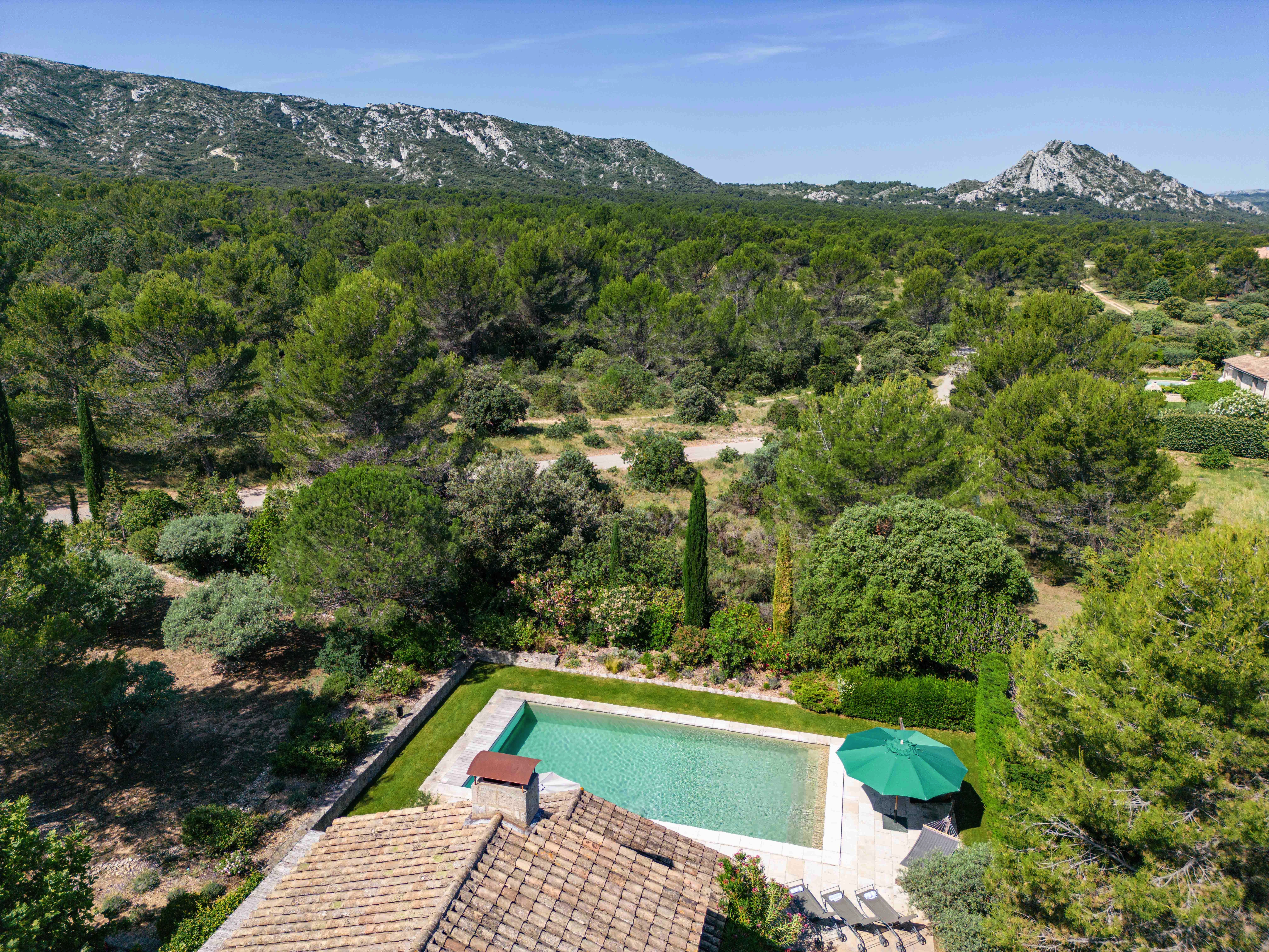 Aerial view of a rectangular swimming pool beside a house, surrounded by lush greenery and distant mountains under a clear blue sky.